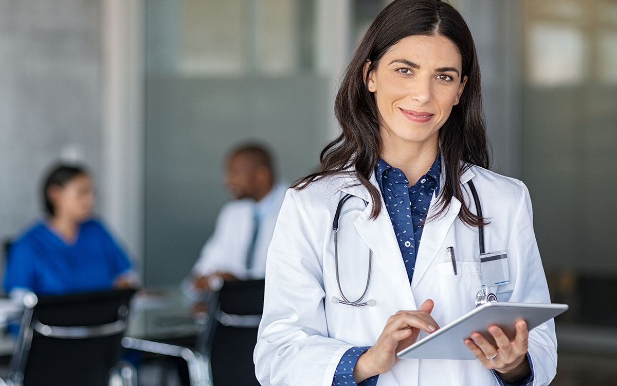 Confident female doctor in a white coat with a stethoscope around her neck, holding a digital tablet